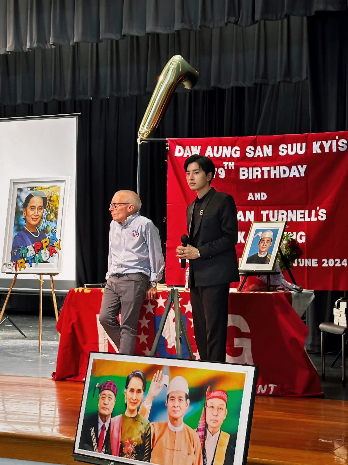 Two men on a stage beside a red banner for Daw Aung San Suu Kyi's birthday, with framed portraits and balloons.