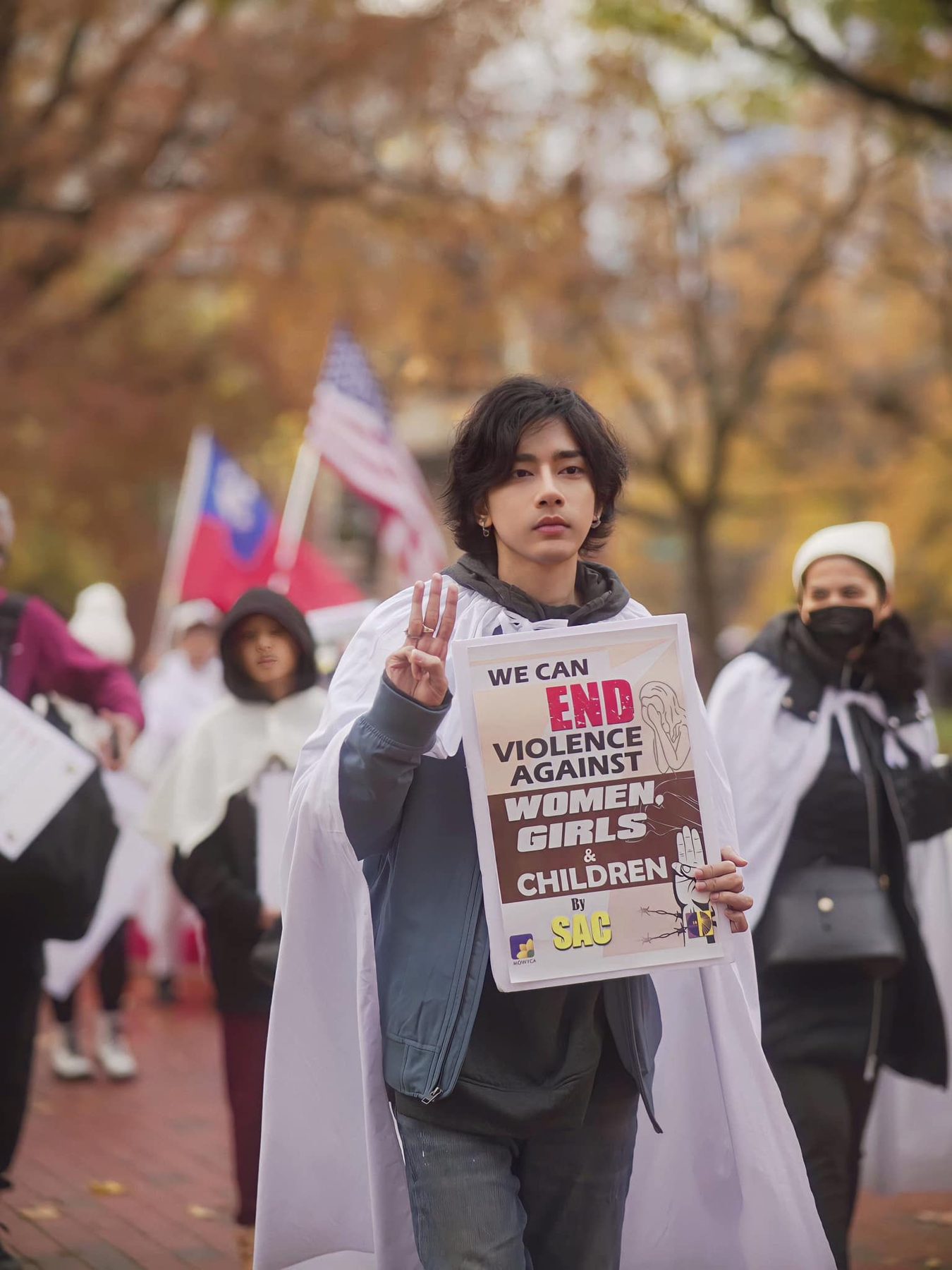 Wai Yan Aung, in a white cape, raises three fingers and holds a sign reading 'We can END violence against women, girls & children' at a march.