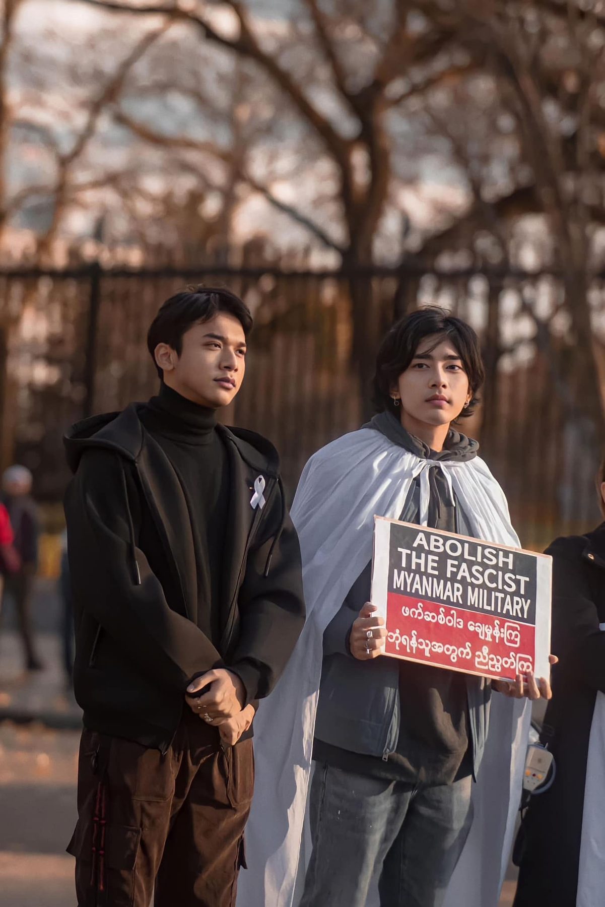 Two young men stand at an evening protest, one holding a sign reading 'Abolish the Fascist Myanmar Military'.