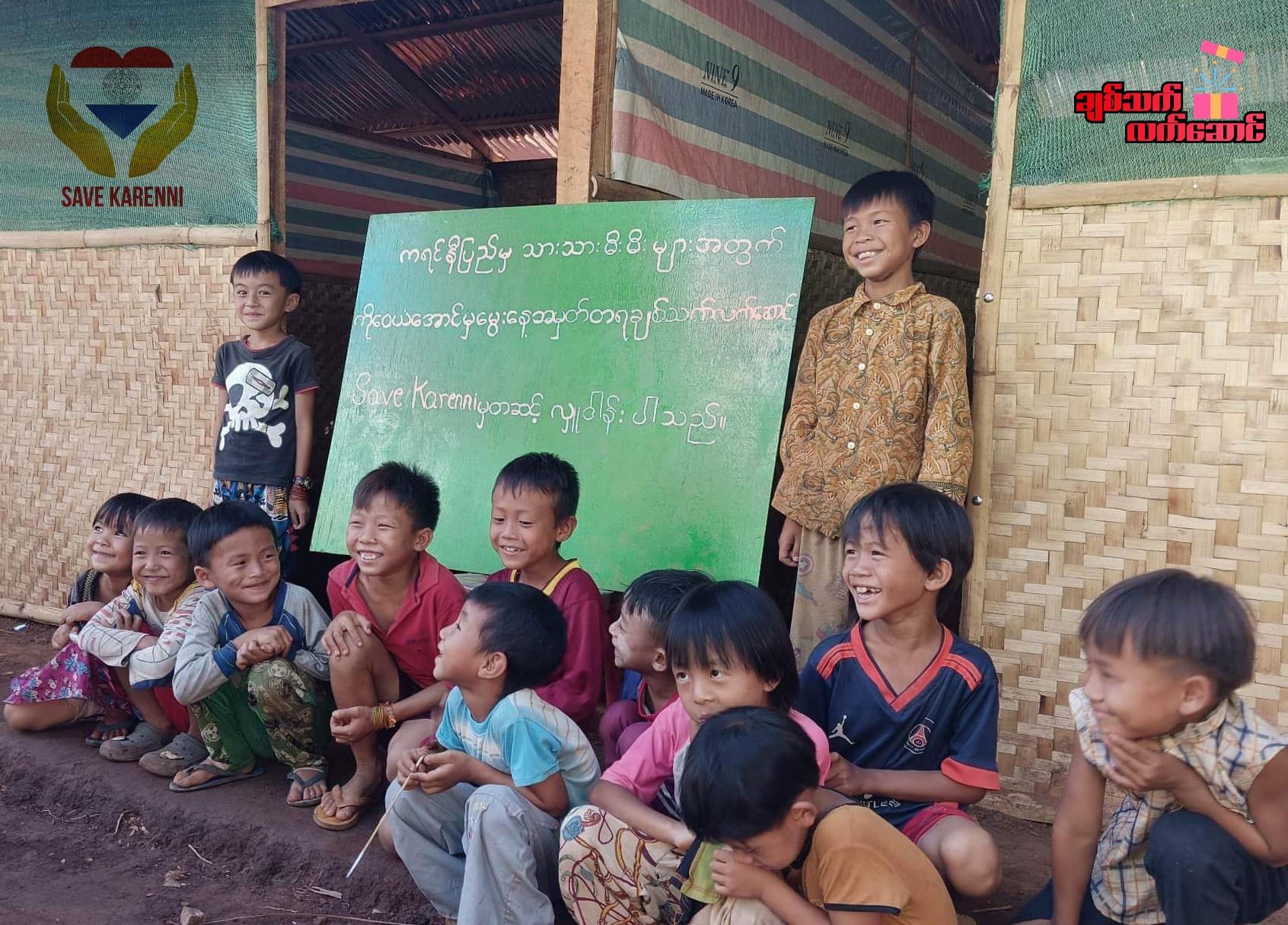 Smiling children crouch in front of a green chalkboard under a Save Karenni banner.