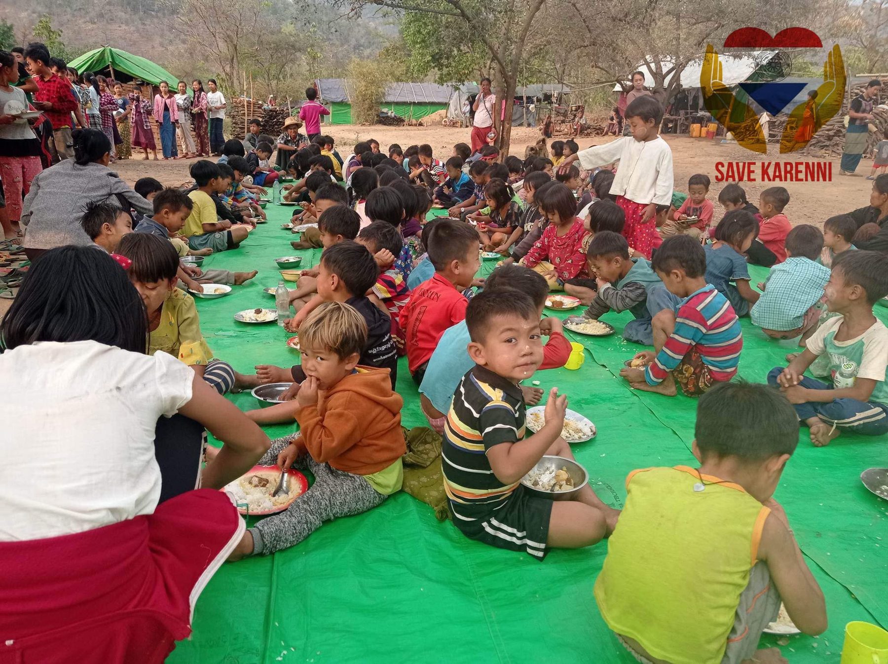 Dozens of children sit on a long green mat outdoors, eating a shared meal, with a Save Karenni logo in the corner.