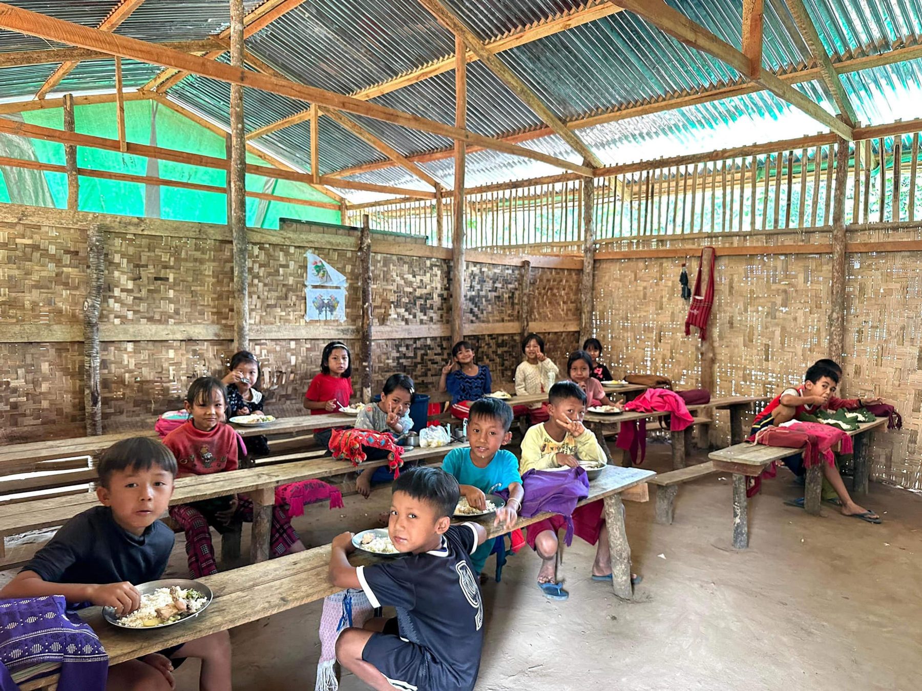 Children eat at wooden benches inside an open-sided classroom with a metal roof.