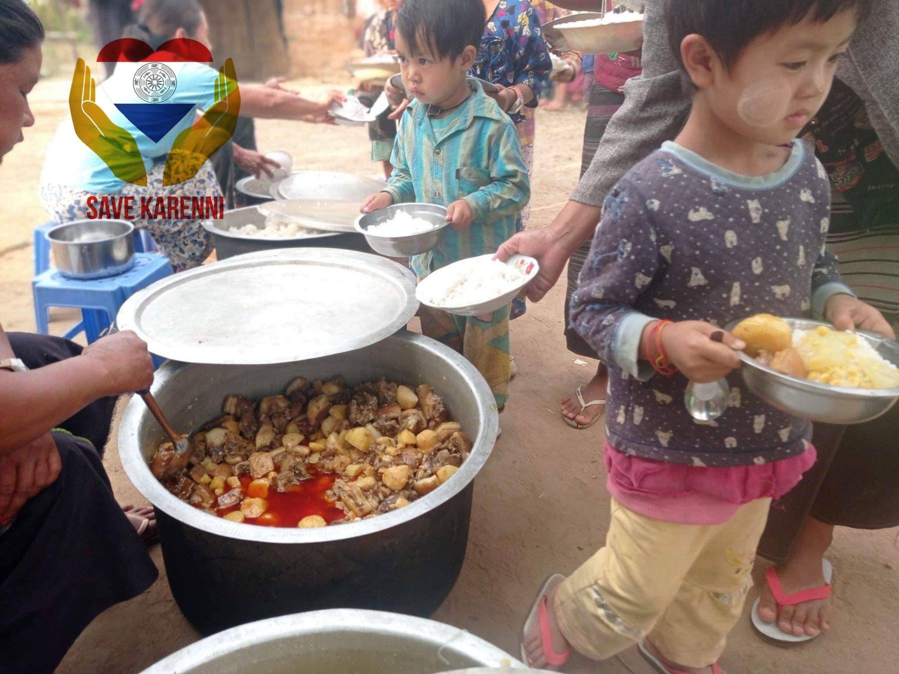 Children with plates wait beside a large pot of curry.