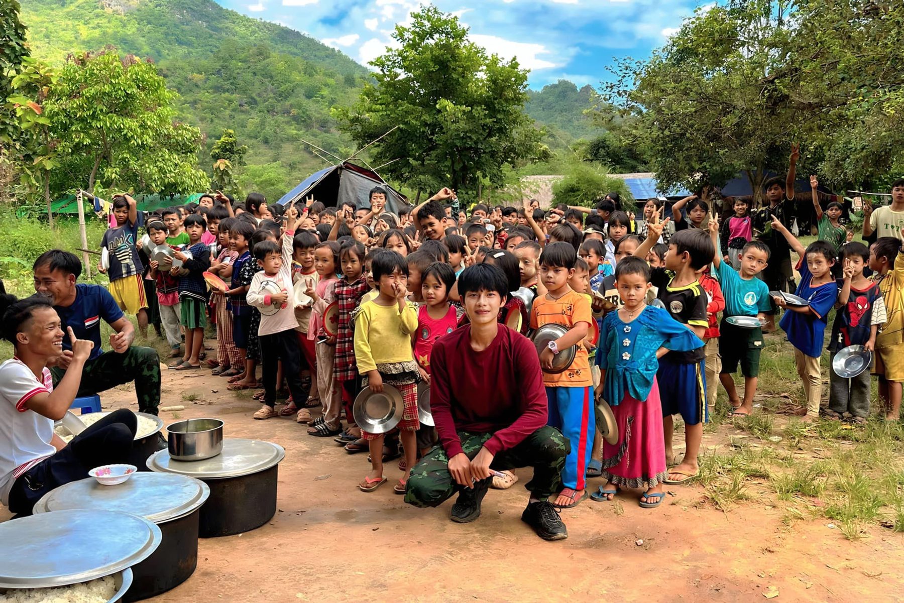 A long line of children with bowls waits beside large cooking pots, with green hills behind.