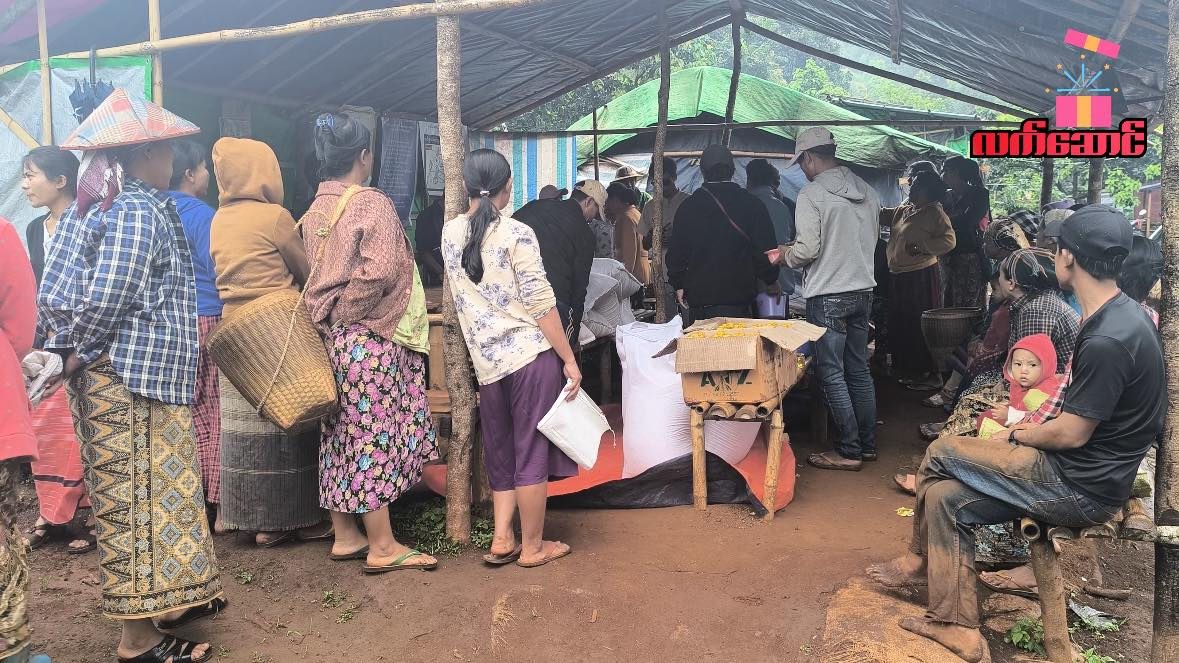People wait under a large blue tarp tent where supplies are handed out.