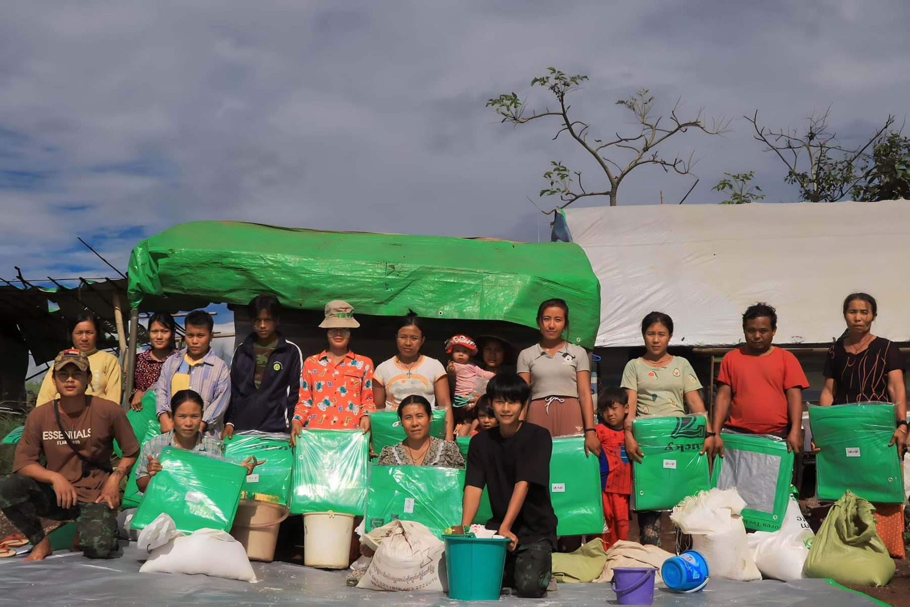 A group of people stand behind green bags of supplies under a green tarp.