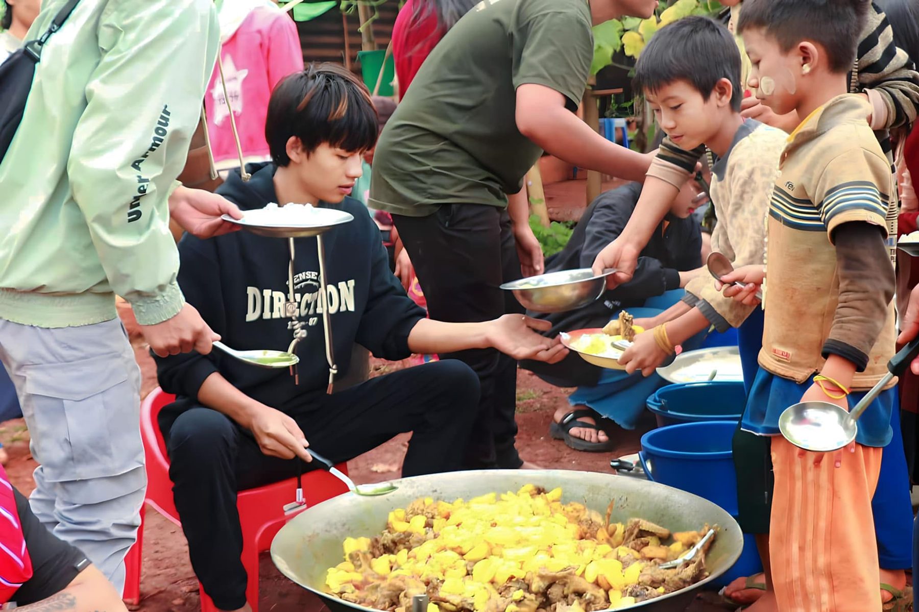 Volunteers serve food from a large pan to children holding plates.