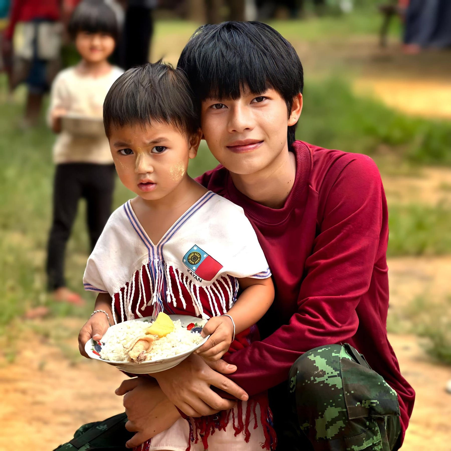 A volunteer in a red sweater hugs a small child who holds a plate of food.