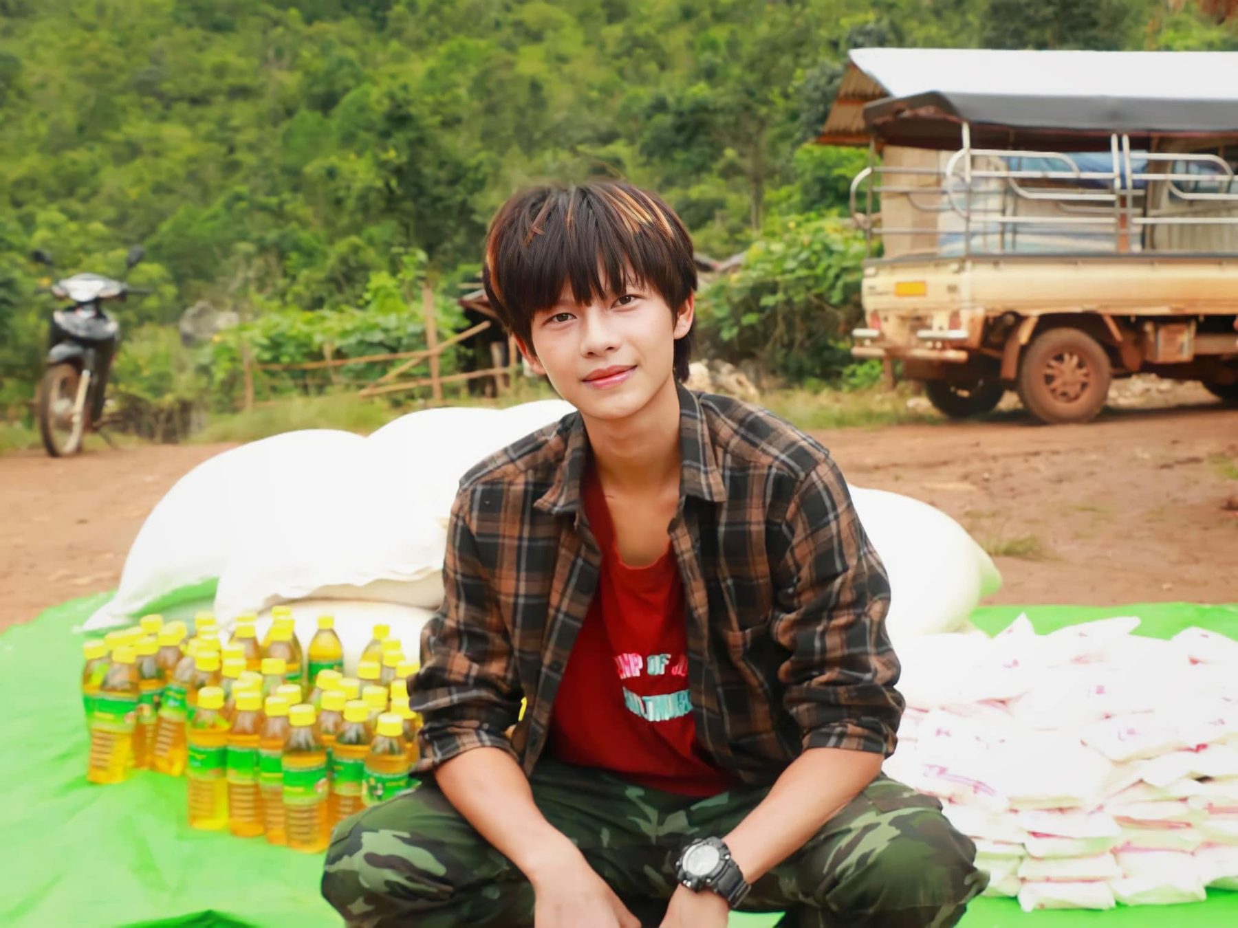 A volunteer in a plaid shirt sits on a green tarp beside bottles of cooking oil and bags of supplies.