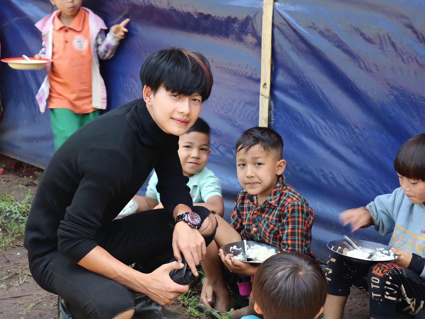 A volunteer in black crouches beside young children eating from plates.